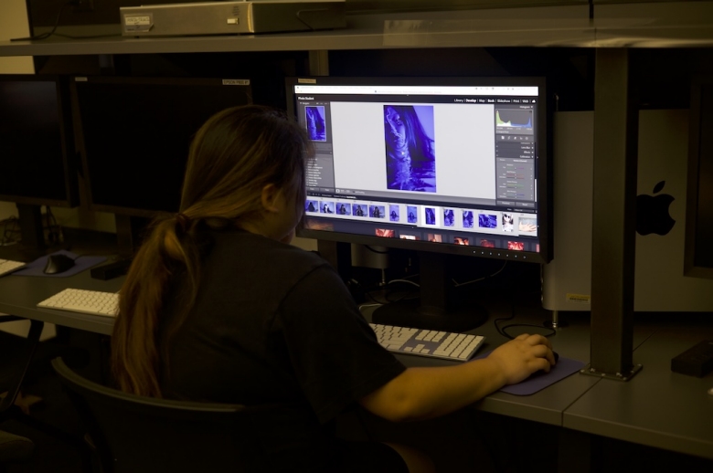 A Tisch Summer High School Photography student sits in front of a computer in a lab editing a blue photo shown on the screen.